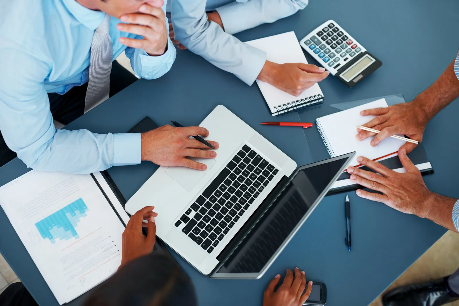 People at a meeting table with a laptop, a calculator and printed charts, seen from above