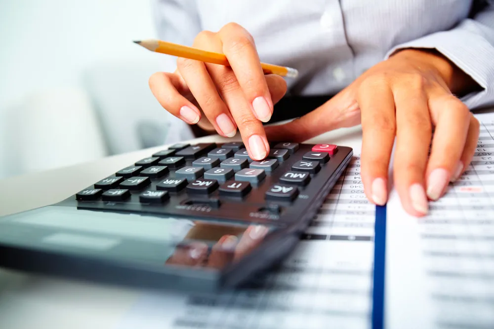 Hands working a desk calculator, holding a pencil, beside a printed spreadsheet