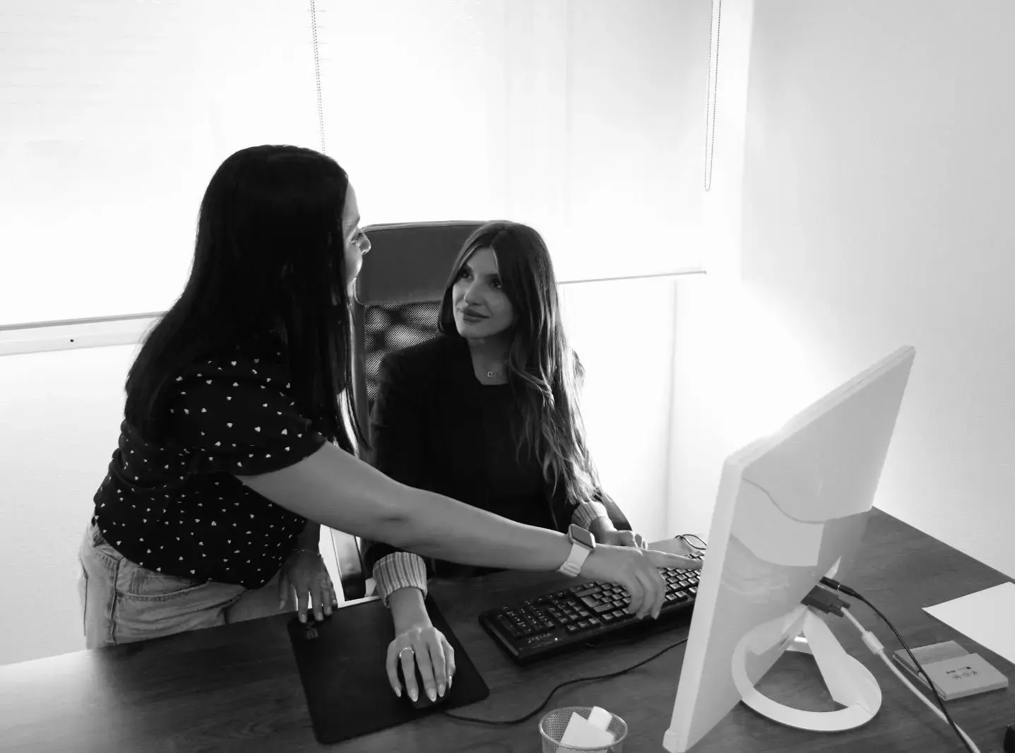 Two colleagues talking at a desk with a desktop computer, in black and white