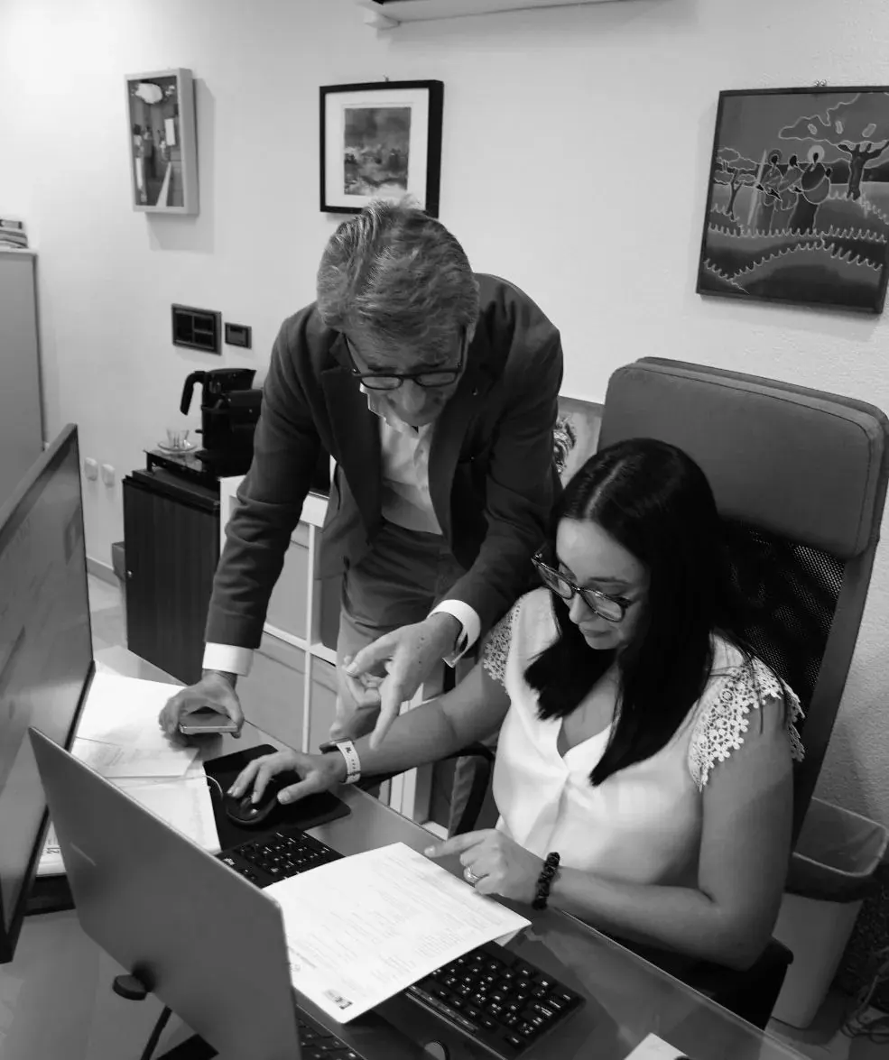 A man in a jacket points at a document while a seated colleague works at a computer, in black and white