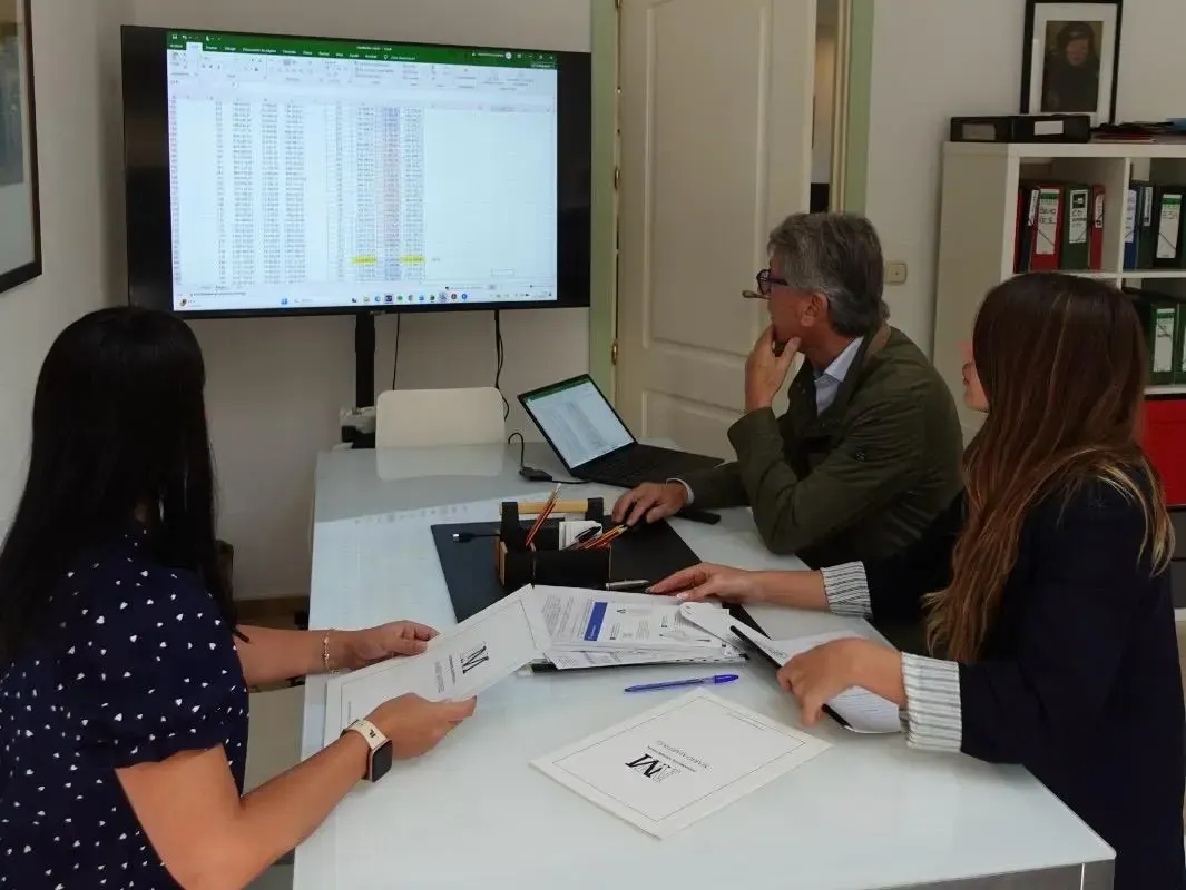 Four people at a meeting table reviewing a spreadsheet shown on a large wall screen