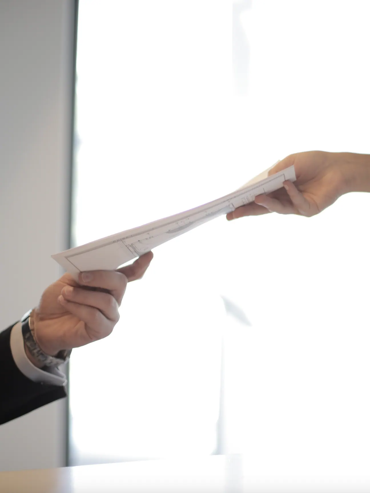 One hand passing a document to another in front of a bright window